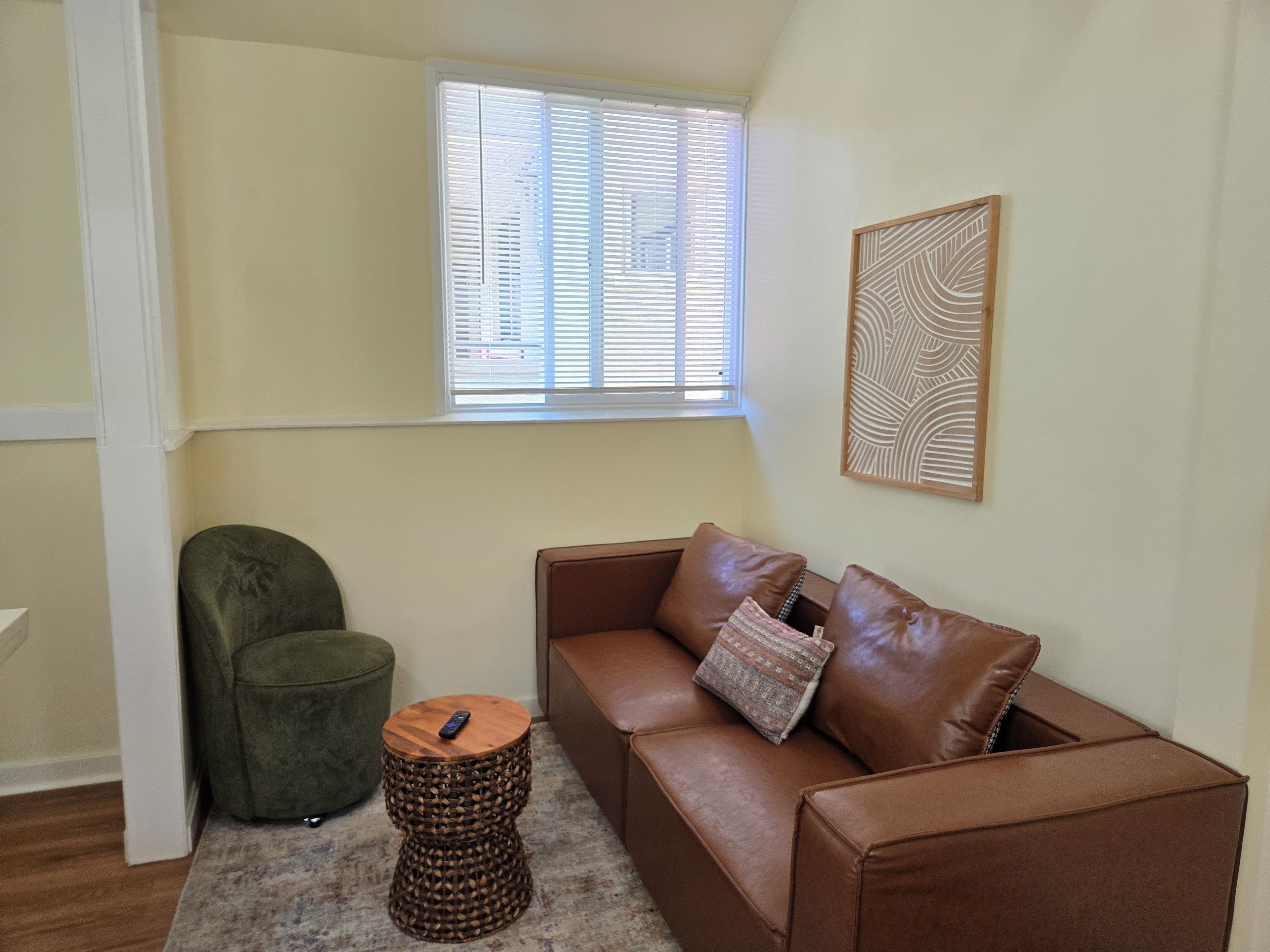 A corner room with a brown leather sofa, a green armchair, a small wooden side table, a woven stool, and a window with blinds.