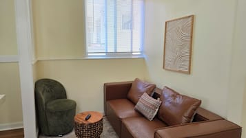 A corner room with a brown leather sofa, a green armchair, a small wooden side table, a woven stool, and a window with blinds.