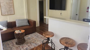 A living room with a brown leather sofa, patterned cushions, a woven side table, and a TV mounted on the wall.