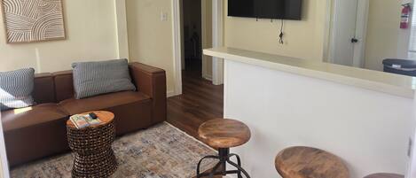 A living room with a brown leather sofa, patterned cushions, a woven side table, and a TV mounted on the wall.
