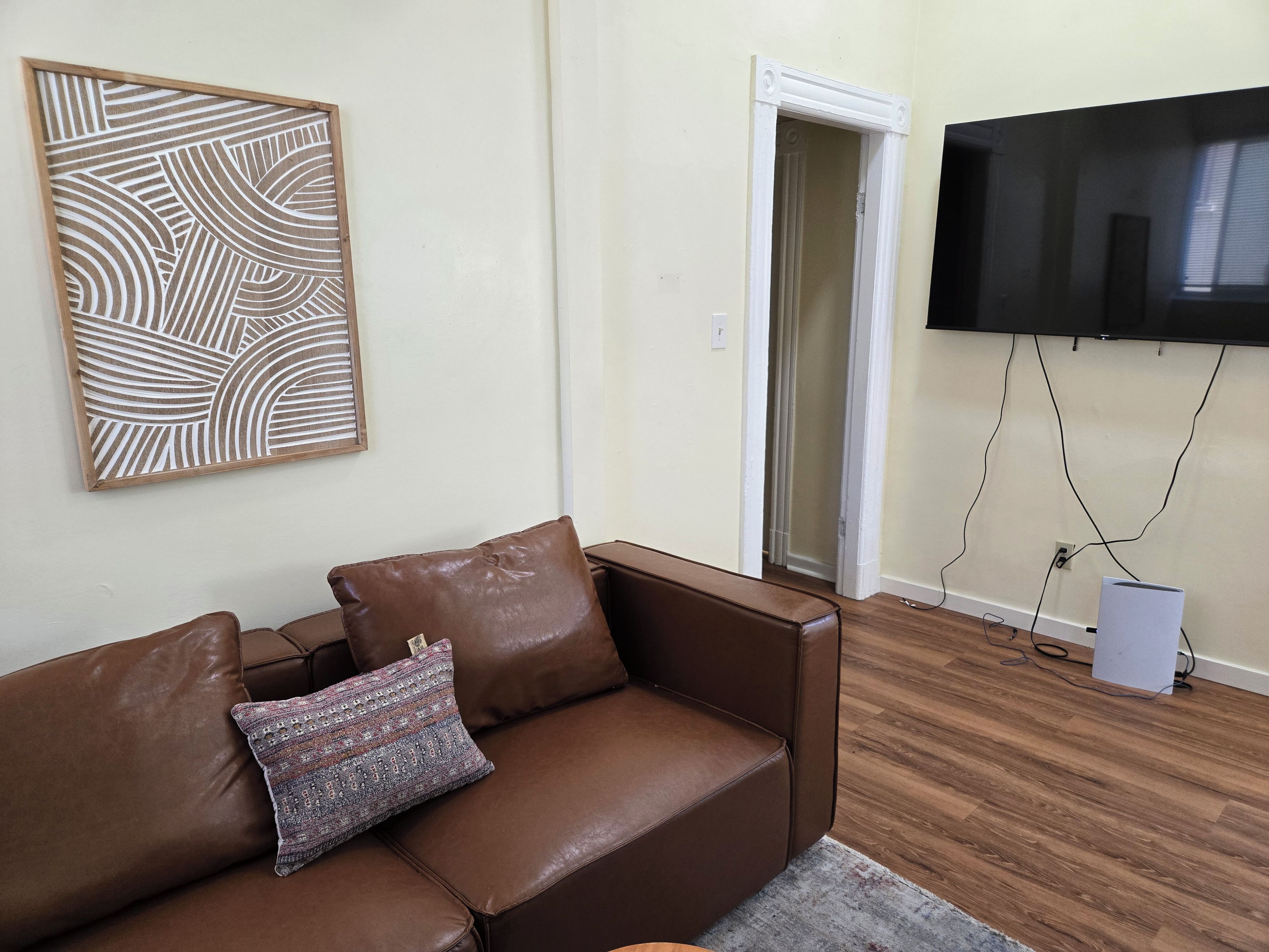 Un sofá de cuero marrón con un cojín estampado, una obra de arte abstracta enmarcada, un televisor montado en la pared y piso de madera.