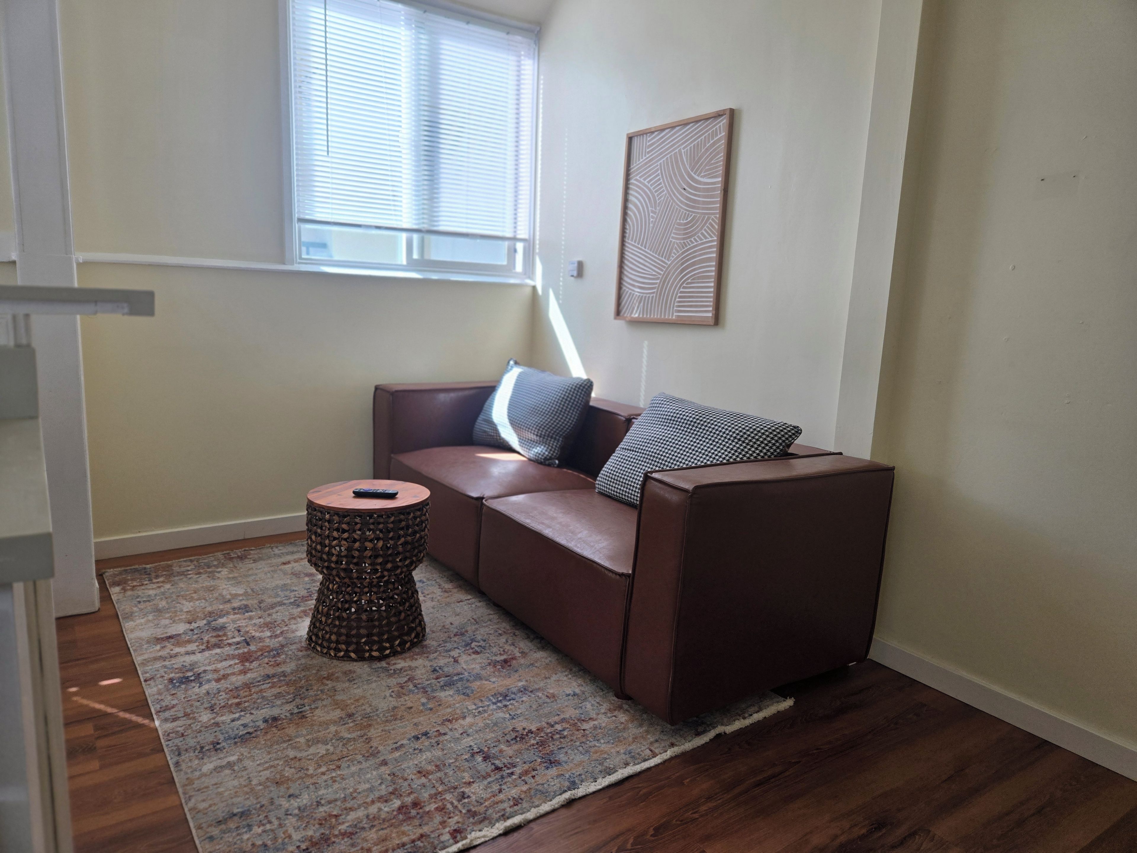 Quarto com sofá de couro marrom, tapete estampado, um banquinho redondo e janela com persianas.