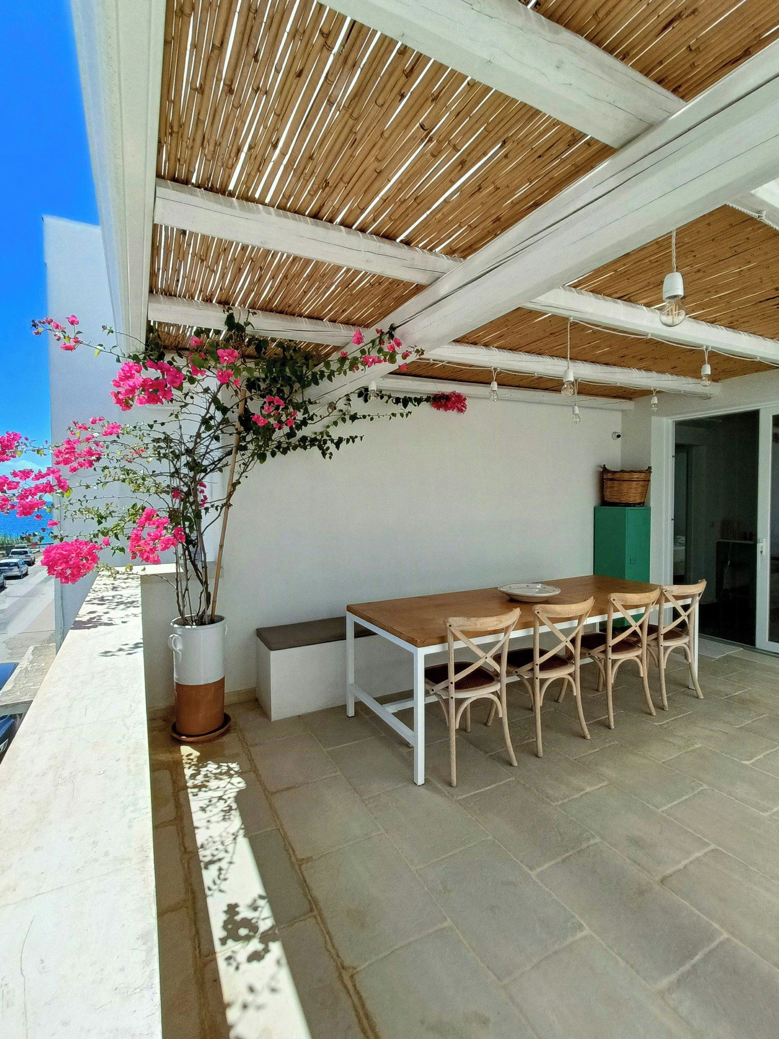 An outdoor patio with a wooden table and chairs, a potted plant with pink flowers, and a view of the sea.