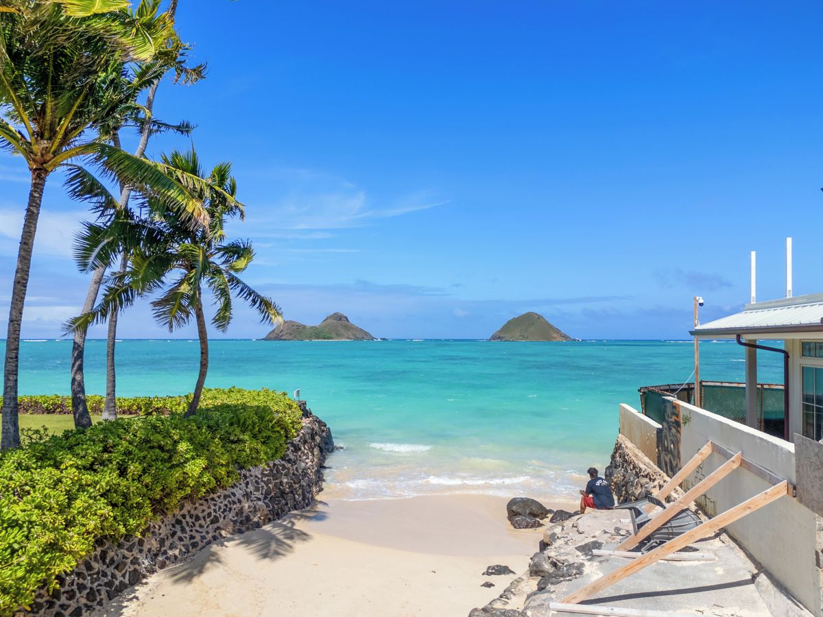 A beach with clear turquoise water, palm trees, and a building with a sloped roof overlooking the ocean.