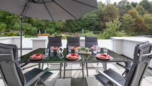 Et udendørs spiseområde med et glasbord, sorte stole og en stor parasol.