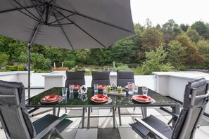 Un espacio de comedor al aire libre con mesa de vidrio, sillas negras y una sombrilla grande.