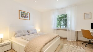 Et moderne soveværelse med seng, sengebord, lampe, vindue med gardiner, skrivebord, stol og et billede i ramme på væggen.