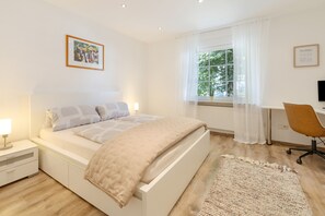Un dormitorio moderno con cama, mesita de noche, lámpara, ventana con cortinas, escritorio, silla y un cuadro en la pared.