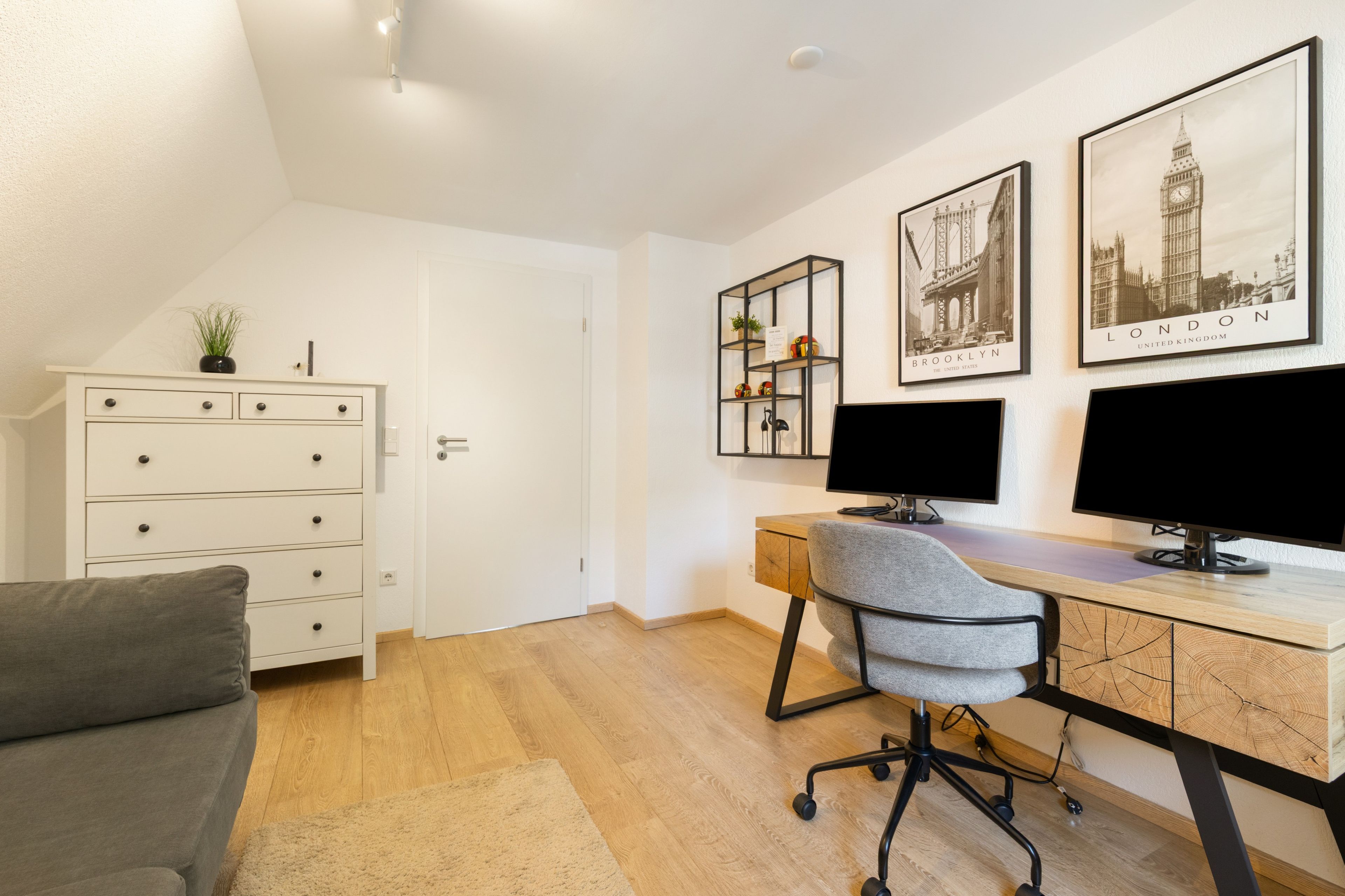 Un espacio de oficina moderno con un escritorio, dos monitores, una silla, un aparador blanco y fotografías enmarcadas de un paisaje urbano en la pared.