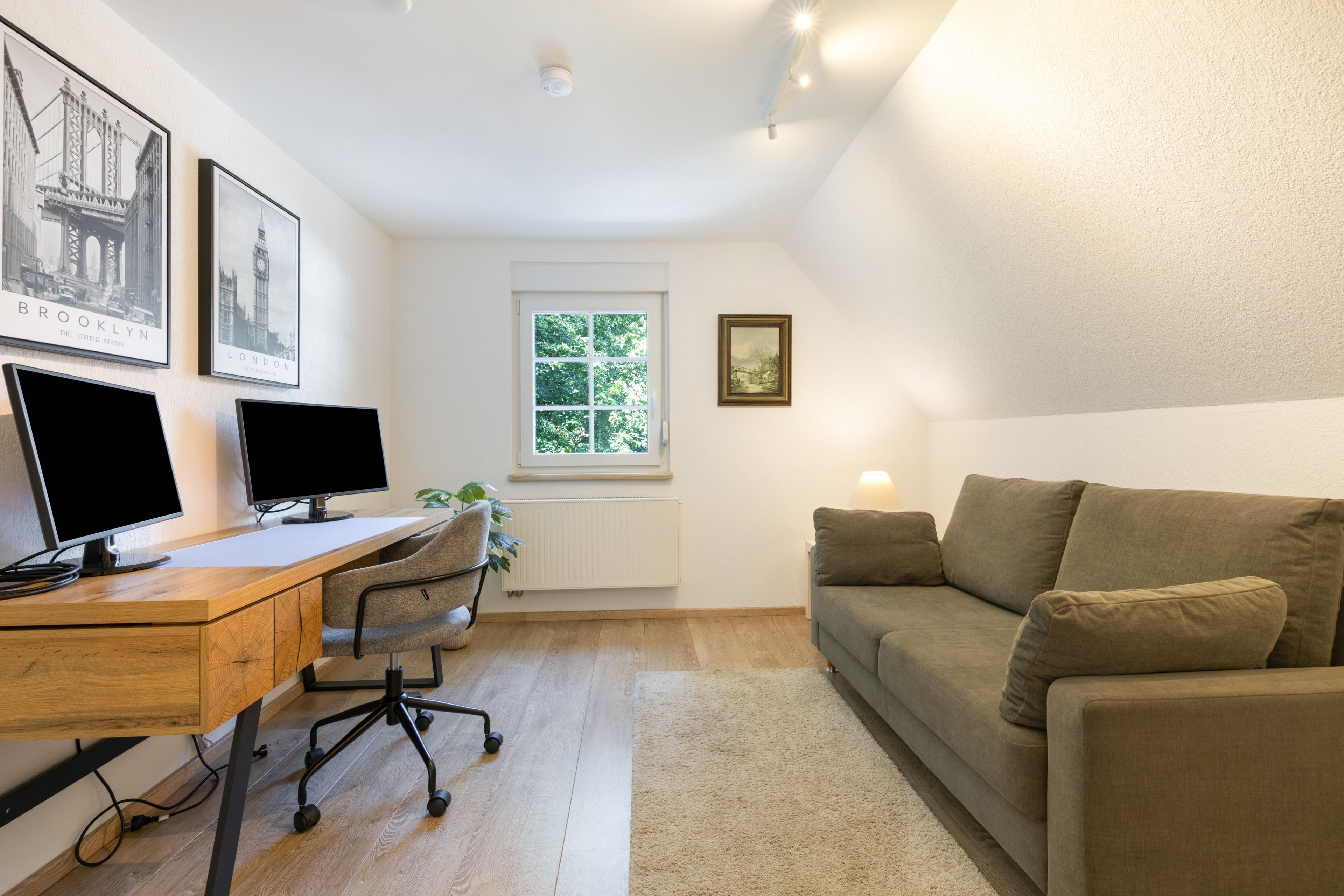 Ein modernes Zimmer mit Schreibtisch, zwei Monitoren, einem Stuhl, einem Fenster mit Blick auf Grünflächen und einer Couch.