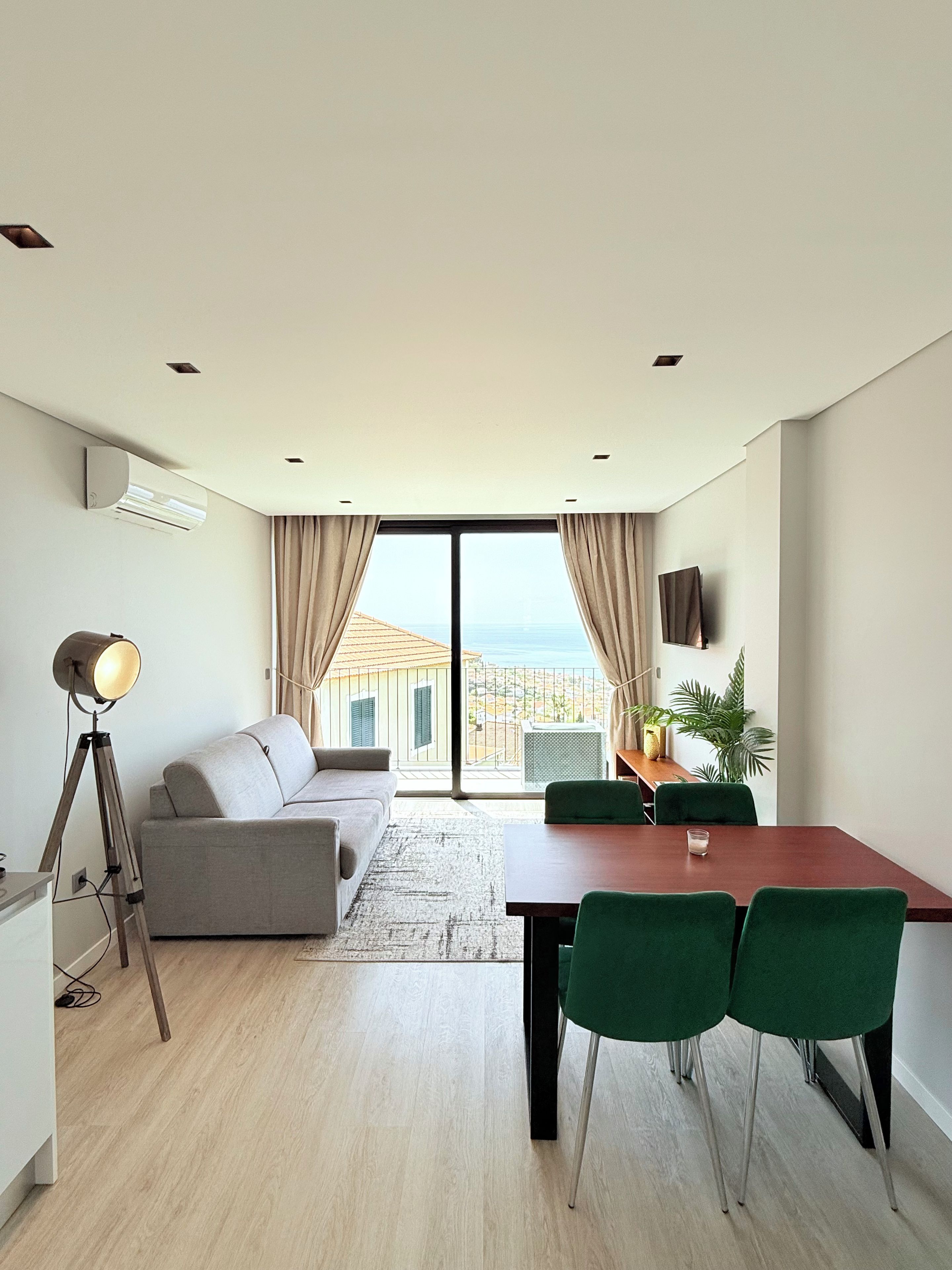 Een moderne woonkamer met een bank, een eettafel met stoelen, een vloerlamp en uitzicht op zee door grote ramen.