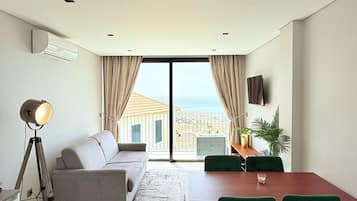 Een moderne woonkamer met een bank, een eettafel met stoelen, een vloerlamp en uitzicht op zee door grote ramen.