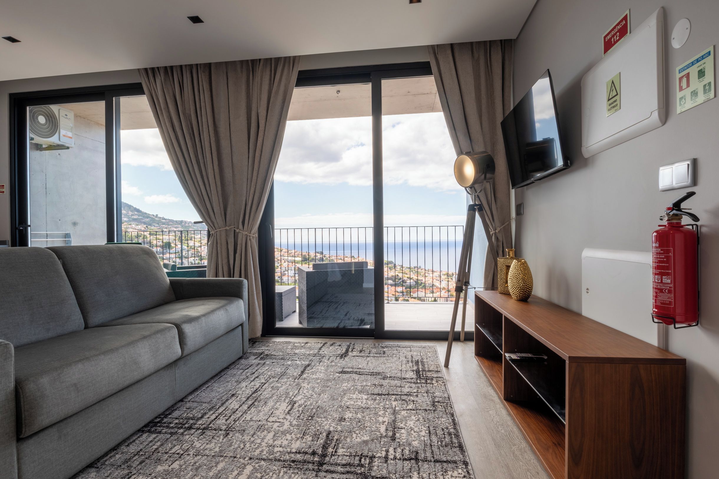 A modern living room with a grey sofa, a flat-screen TV mounted on the wall, a fire extinguisher, and a view of the city through large windows.