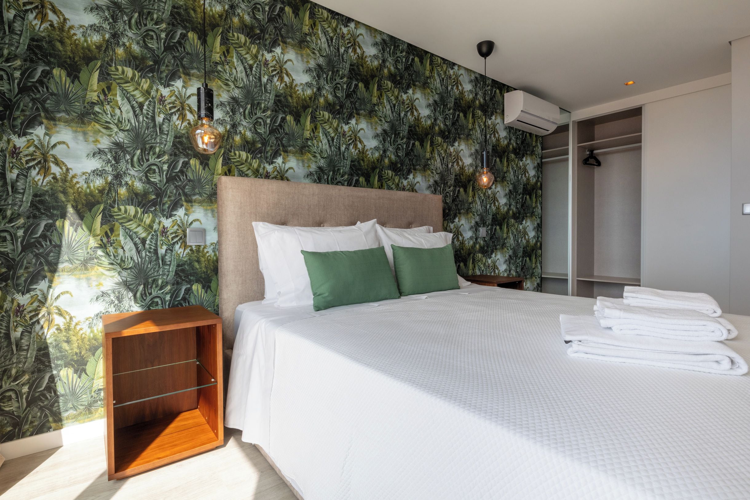 A hotel room with a bed, a nightstand, a wooden side table, a wardrobe, and a wall with a tropical leaf pattern.