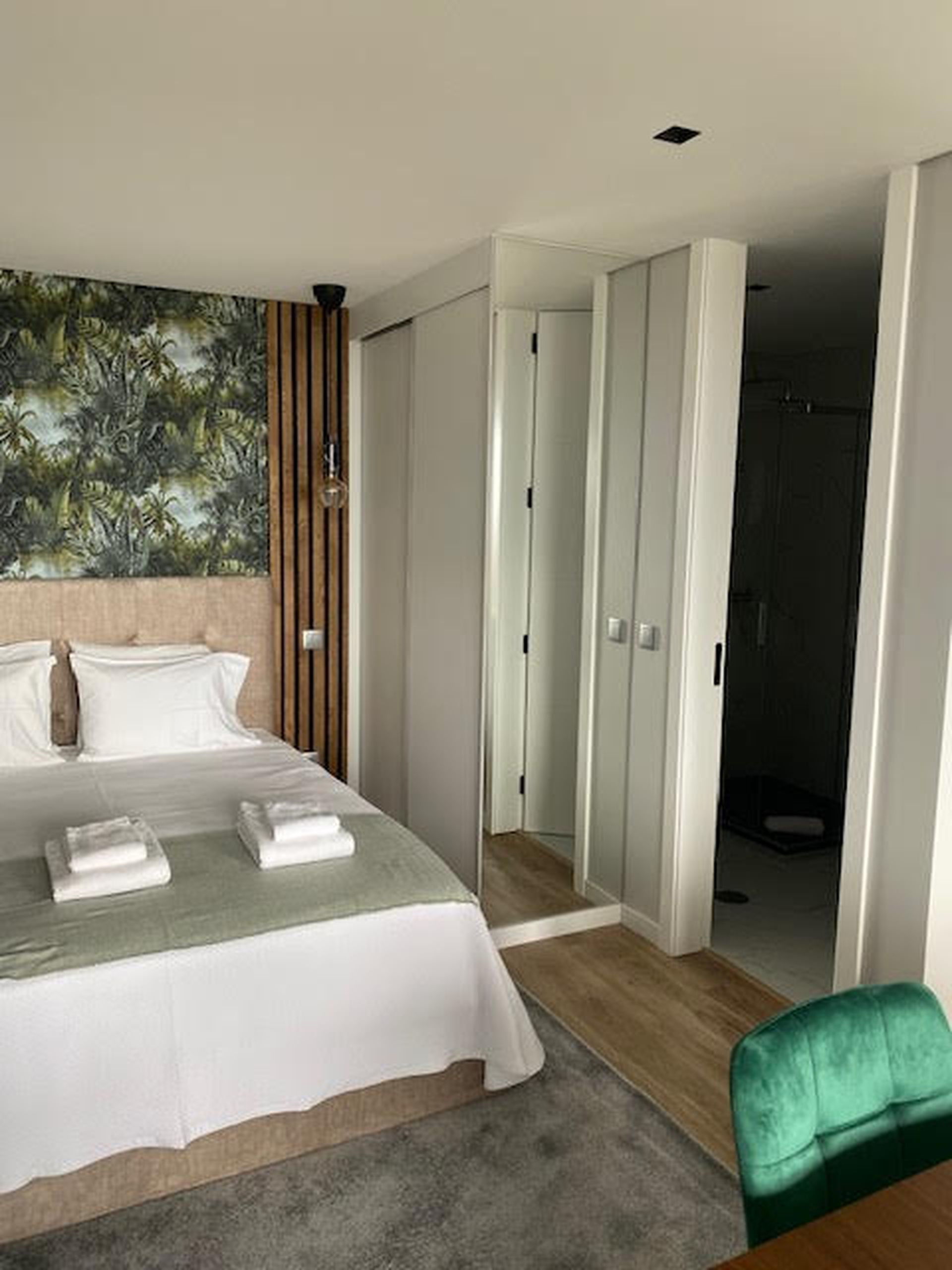 A hotel room with a bed, a green chair, a wardrobe, and a door leading to a bathroom.