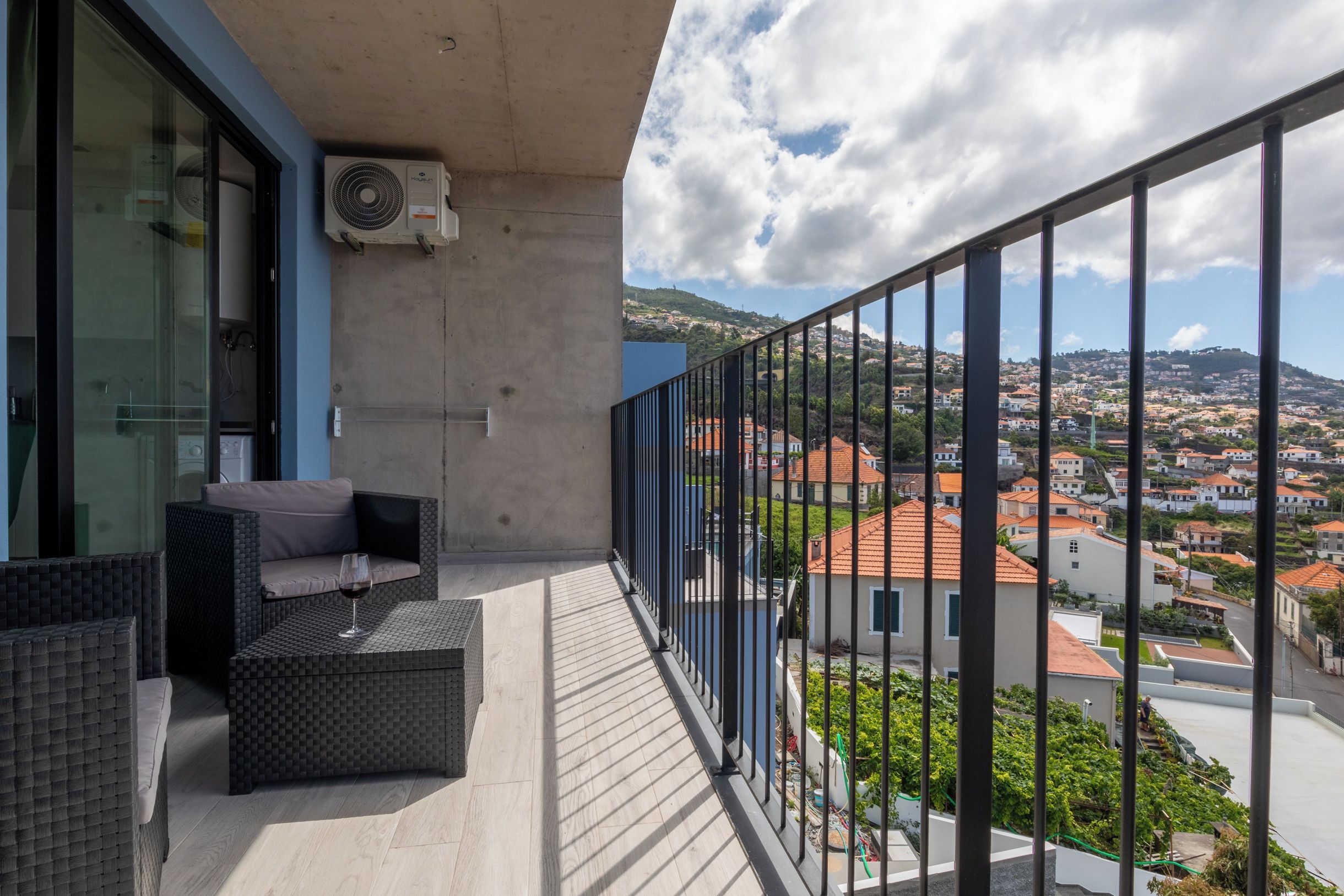A balcony with a wicker sofa and a glass table, overlooking a town with red-roofed buildings.