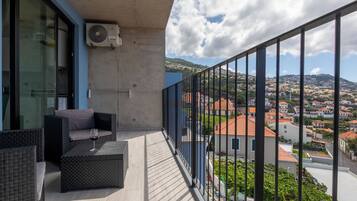 A balcony with a wicker sofa and a glass table, overlooking a town with red-roofed buildings.