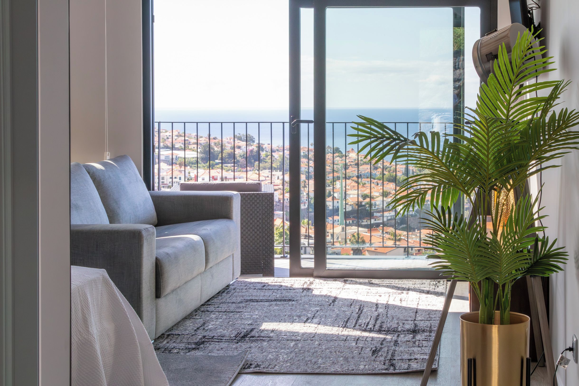 Una sala de estar con un sofá gris, un tapete estampado, una planta grande en maceta y vistas a un pueblo costero desde un balcón.