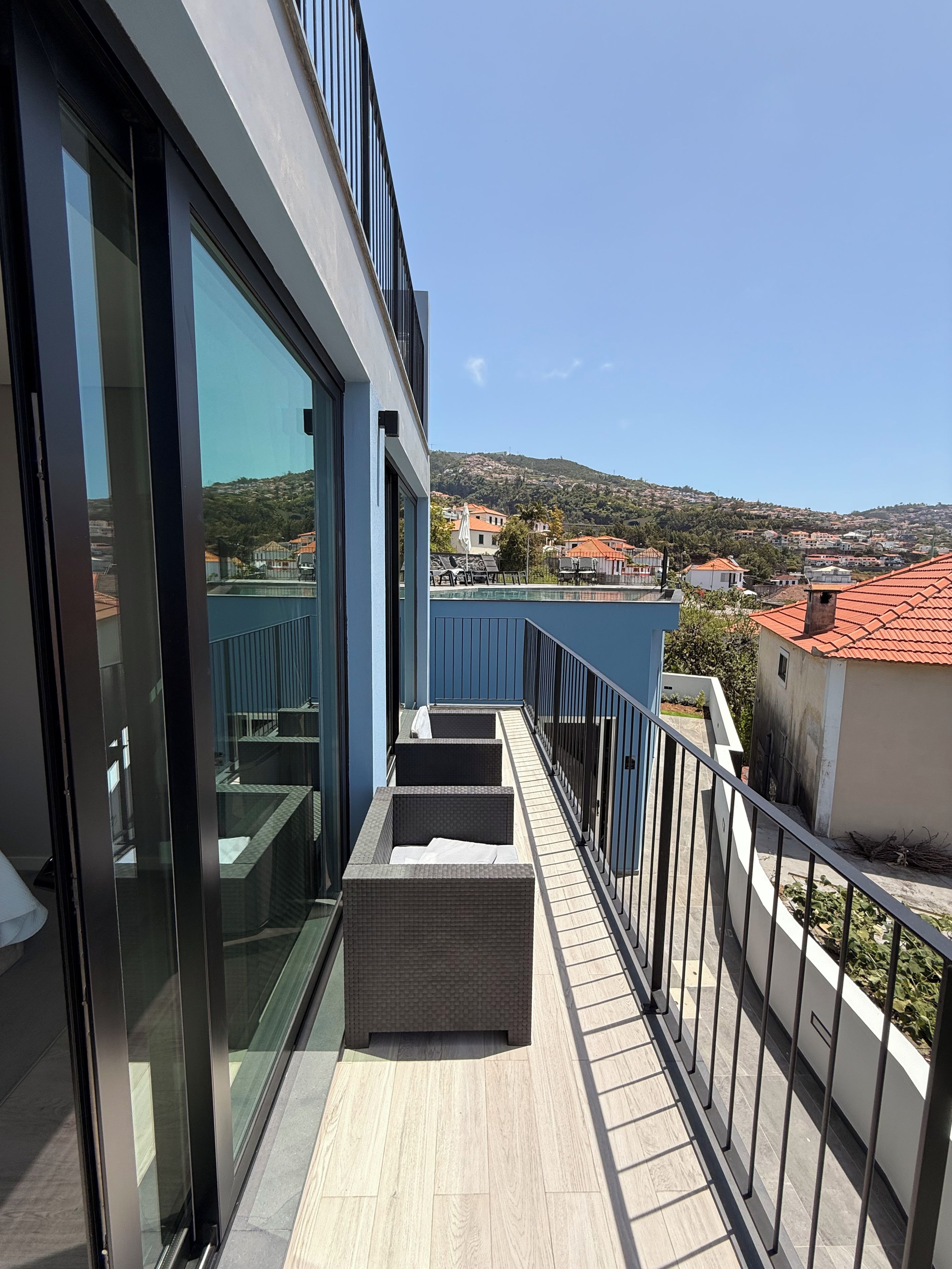 A balcony with a view of a hillside town, a pool, and a building with a red roof.