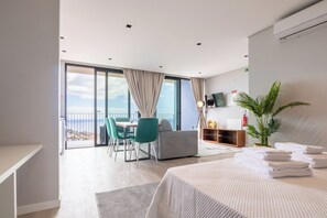 A modern hotel room with a balcony, a bed, a desk, and a seating area with a view of the sea.