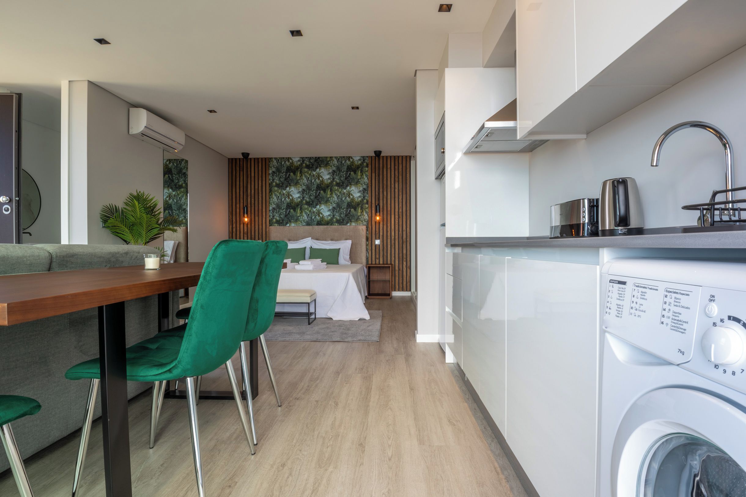 A modern kitchen with a washing machine, a dining area with a green chair, and a bedroom with a bed and curtains.