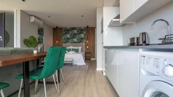 A modern kitchen with a washing machine, a dining area with a green chair, and a bedroom with a bed and curtains.