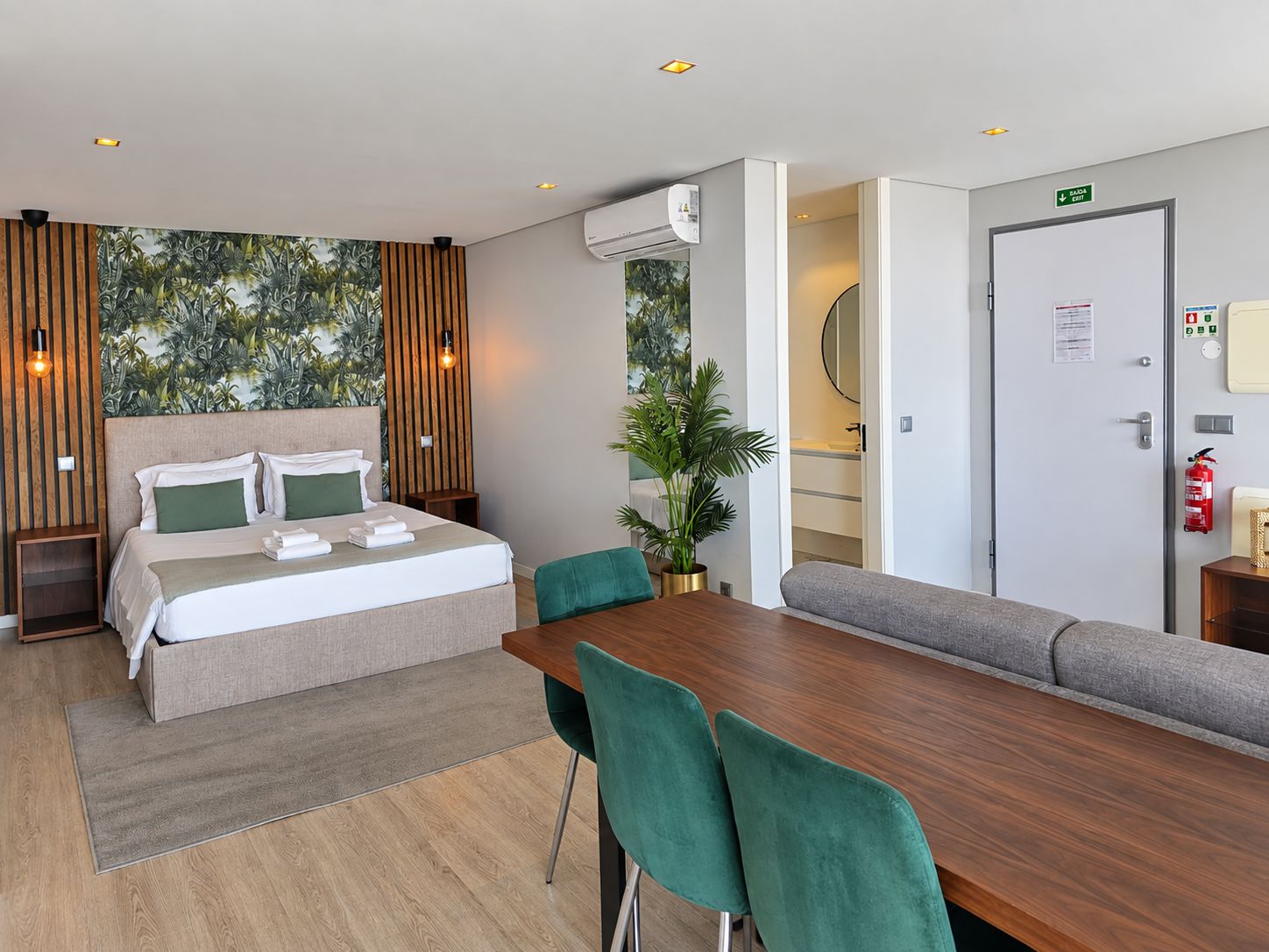 A modern hotel room with a bed, a dining table, chairs, a sofa, a potted plant, and an air conditioning unit.