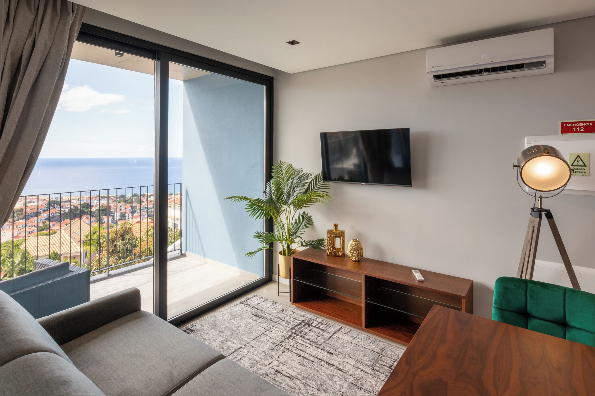 A modern living room with a large sliding glass door leading to a balcony, a flat-screen TV mounted on the wall, a wooden TV stand with decorative items, and a floor lamp.