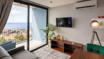A modern living room with a large sliding glass door leading to a balcony, a flat-screen TV mounted on the wall, a wooden TV stand with decorative items, and a floor lamp.