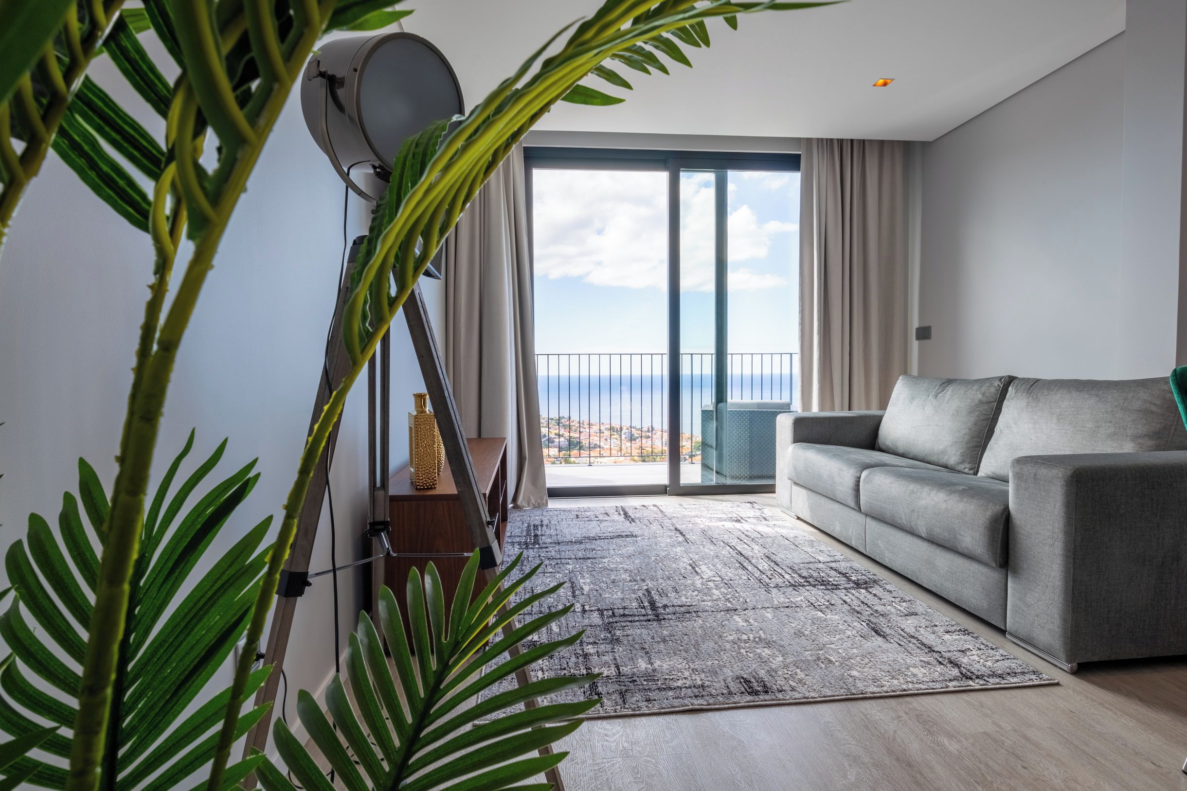 Un salón moderno con un sofá gris, un ventanal con vistas y una planta en primer plano.