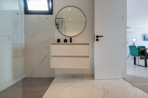 A modern bathroom with a marble floor, a white vanity with a round mirror, and a door leading to a dining area.