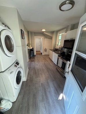 Een wasruimte met een wasmachine en droger, een keuken met een fornuis en spoelbak, en een gang naar een deur.
