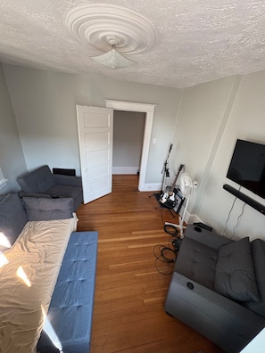 Een kamer met een bed, twee slaapbanken, een gitaar, een ventilator en een televisie.