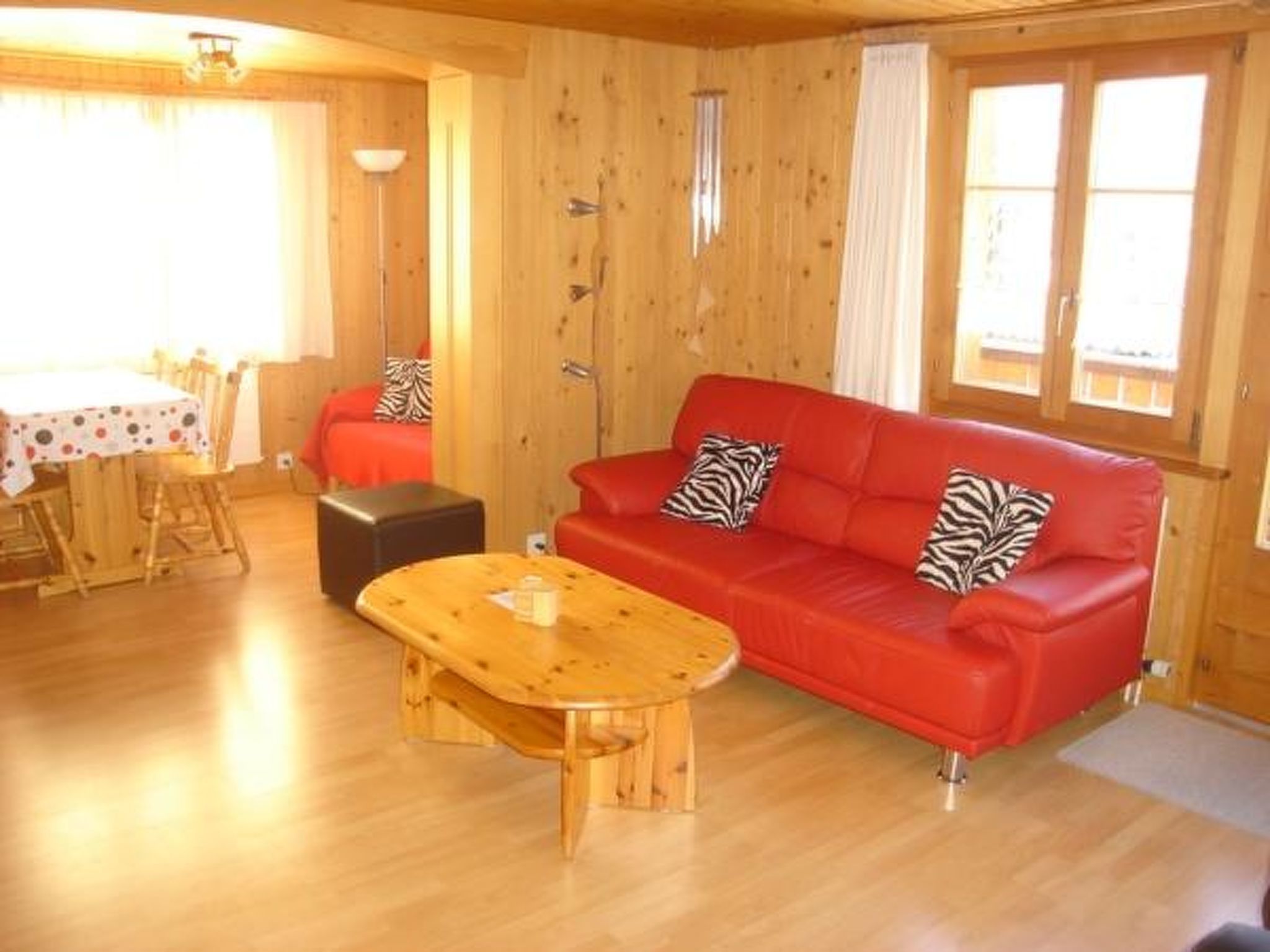A wooden interior with a red sofa, zebra-patterned pillows, a wooden coffee table, and a dining area with a table and chairs.