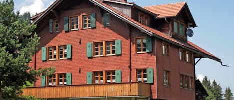 Un grand bâtiment à deux étages, doté d’un toit à forte pente, de nombreuses fenêtres avec volets et d’un balcon en bois.