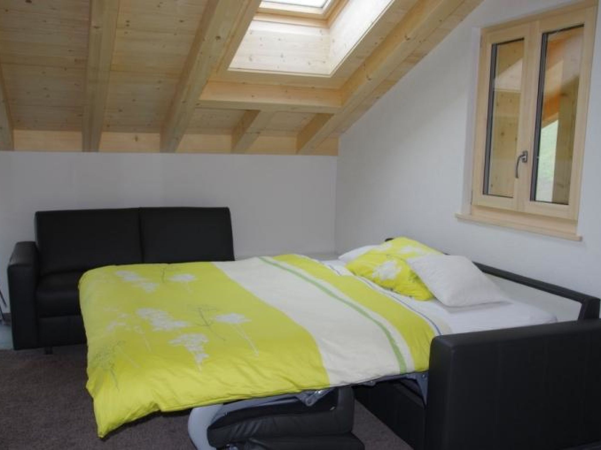 A bedroom with a skylight, a bed with a yellow and white bedspread, a black chair, and a window with wooden shutters.