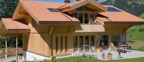 Une maison en bois dotée d’une grande véranda, d’un coin repas avec du mobilier d’extérieur, et offrant une vue sur des collines verdoyantes en arrière-plan.