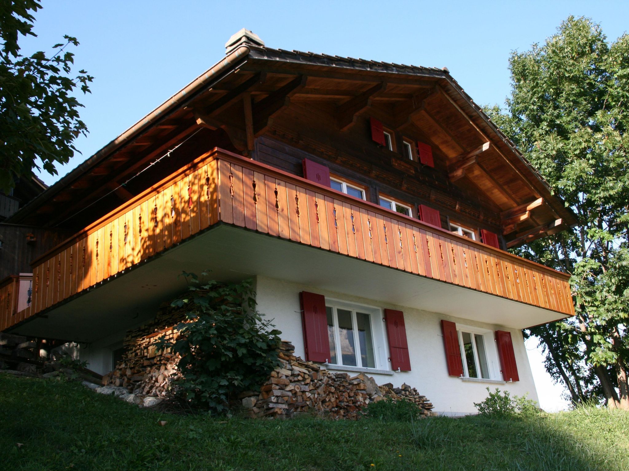 A two-story wooden house with a balcony, red shutters, and a stone foundation.
