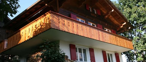 Une maison en bois à deux étages, dotée d’un balcon, de volets rouges et de fondations en pierre.