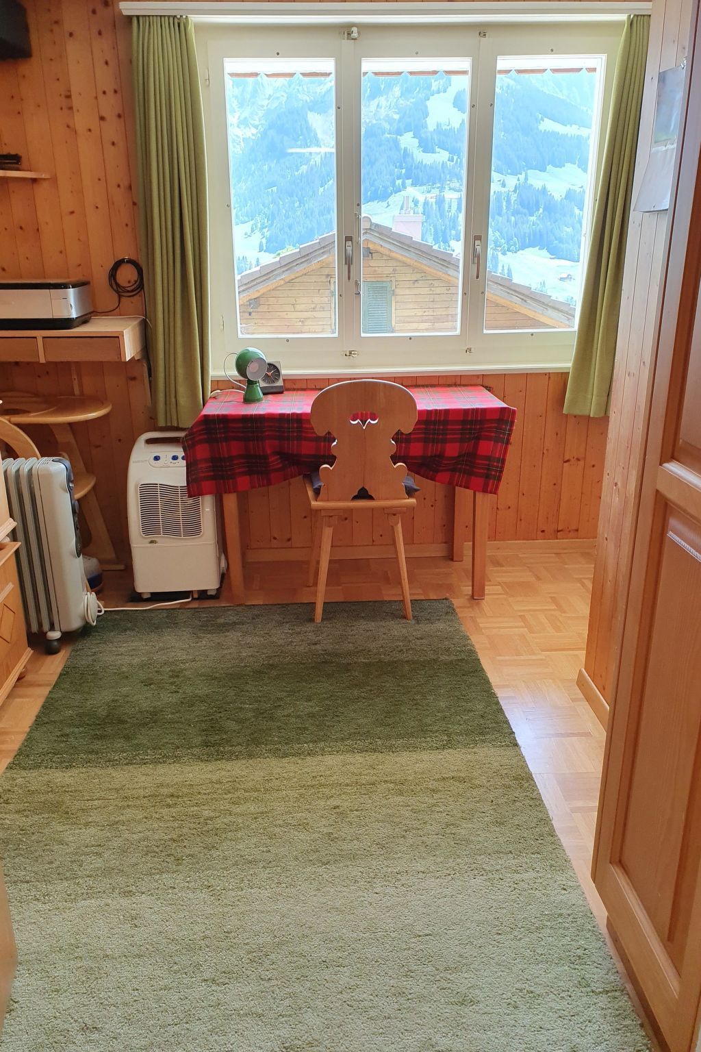 Une pièce au décor intérieur en bois, une table recouverte d’une nappe à carreaux, une chaise, un radiateur et une fenêtre donnant sur un paysage montagneux.