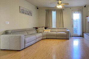 Ein Wohnzimmer mit einem beigefarbenen Ecksofa, einem Deckenventilator, einem Fenster mit Vorhängen und einem Holzboden.