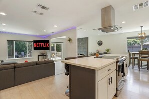 A modern kitchen with a central island, a dining area with a table and chairs, and a living area with a sofa and a TV displaying 'NETFLIX'. - Clean Modern Living | College-Glen Gem Near River (Sacramento)