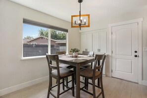 A dining area with a table and chairs, a hanging light fixture, a window with a view of a fence and houses, and a white door. - Clean Modern Living | College-Glen Gem Near River (Sacramento)