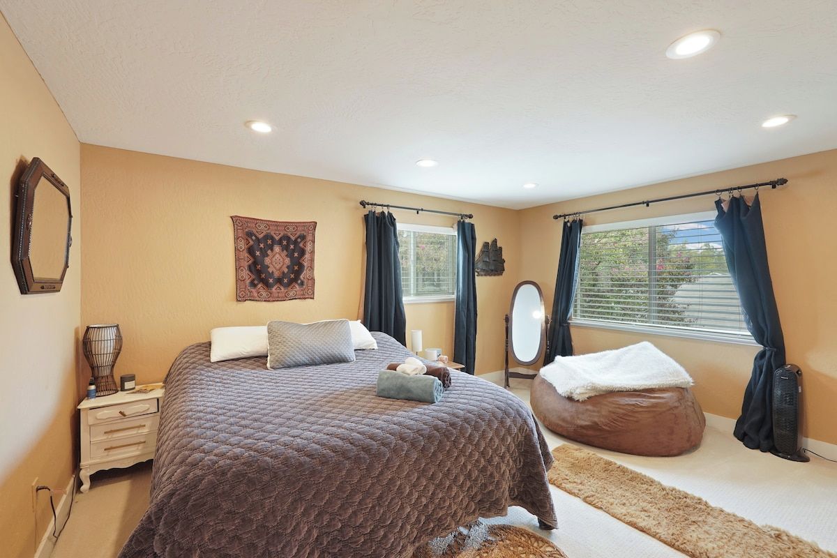 A bedroom with a large bed, a bedside table, a mirror, and a window with curtains.