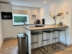 A modern kitchen with a central island, bar stools, a coffee machine, and a trash bin.
