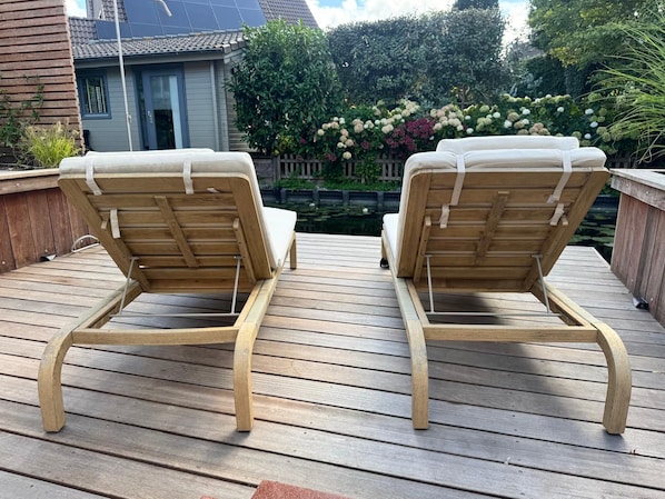 Two wooden lounge chairs with cushions on a wooden deck, a house in the background, and a garden with various plants and flowers.