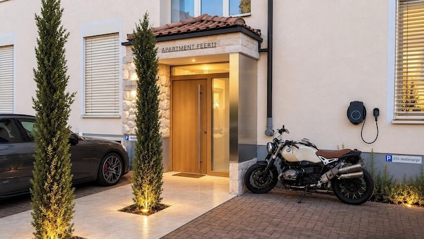 Ein Gebäudeeingang mit einem Schild, auf dem 'APARTMENT FEER11' steht, einem abgestellten Motorrad und einem Auto.