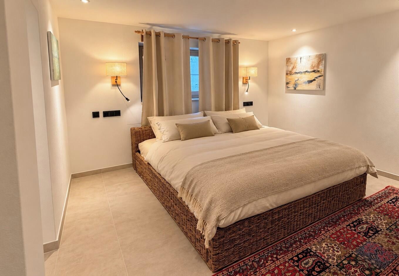 Une chambre à coucher avec un grand lit, deux lampes fixées au mur, une fenêtre avec des rideaux et un tapis à motifs.