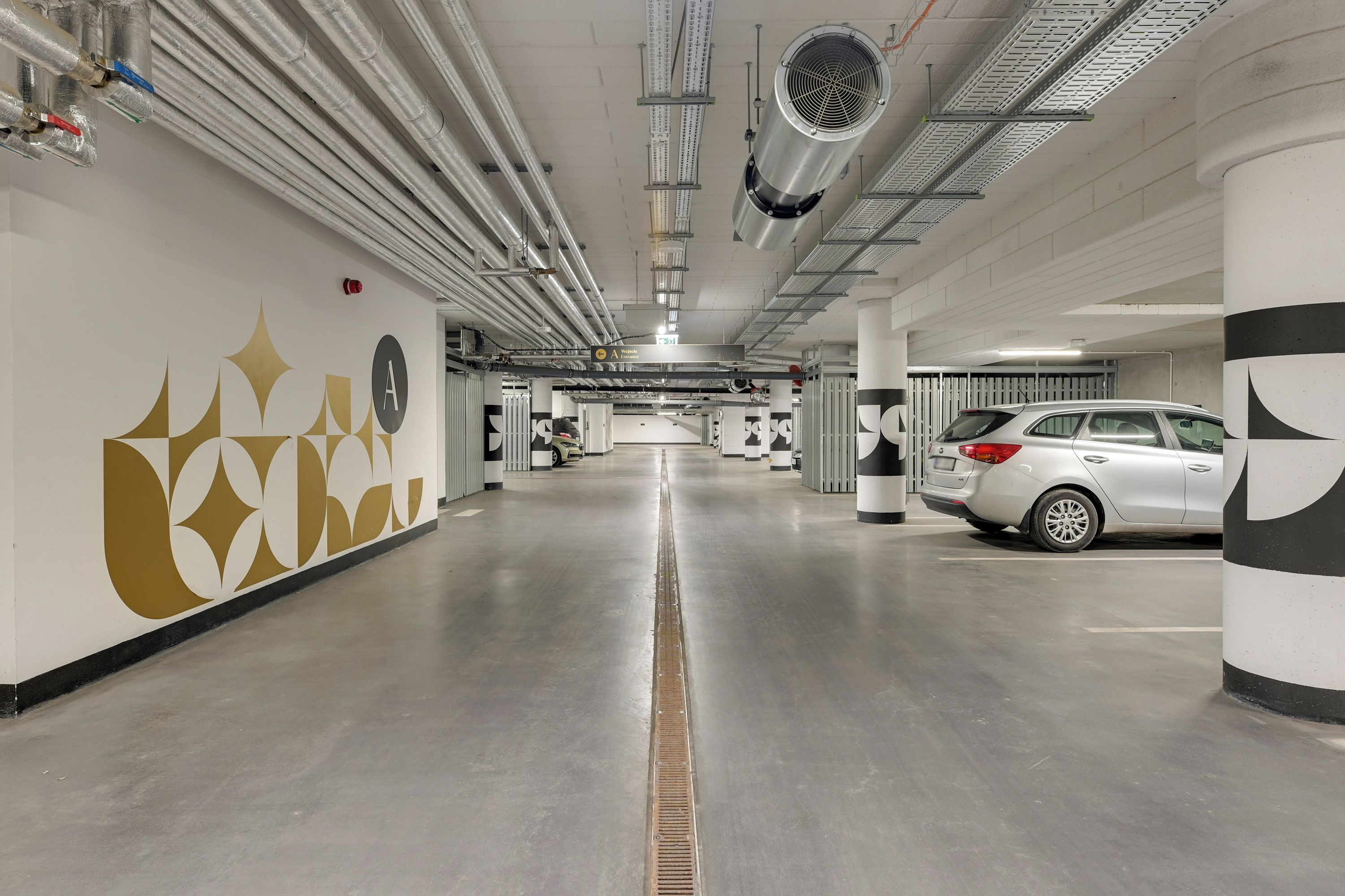 A parking garage with a white car parked on the right, a ventilation system on the ceiling, and a wall with geometric patterns.