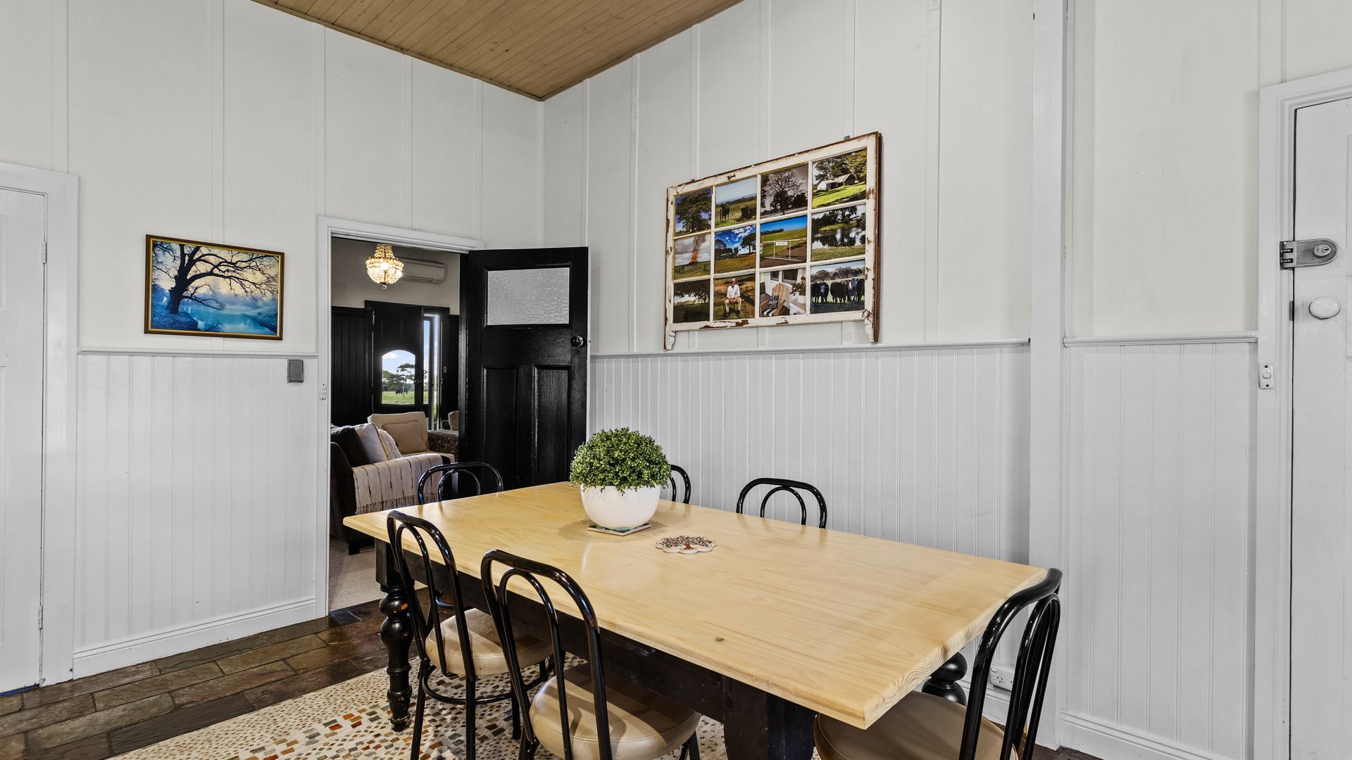A dining area with a wooden table, chairs, a potted plant, and a wall with framed pictures.